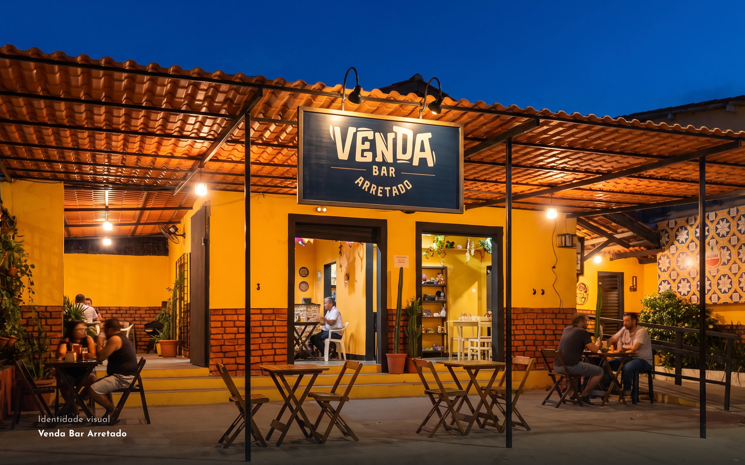 Fachada amarela do Venda Bar Arretado à noite, com a placa sob o pergolado e mesas de madeira
